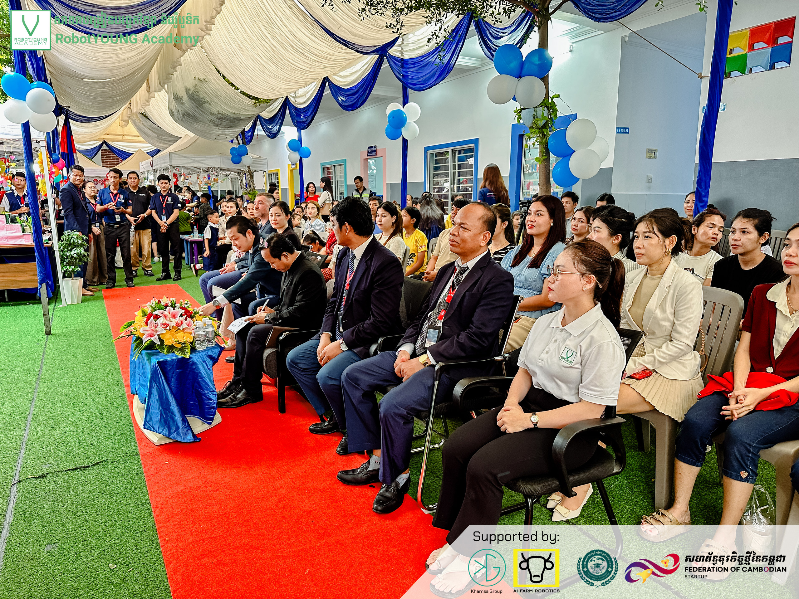 An audience seated beside a red carpet under blue and white event decorations