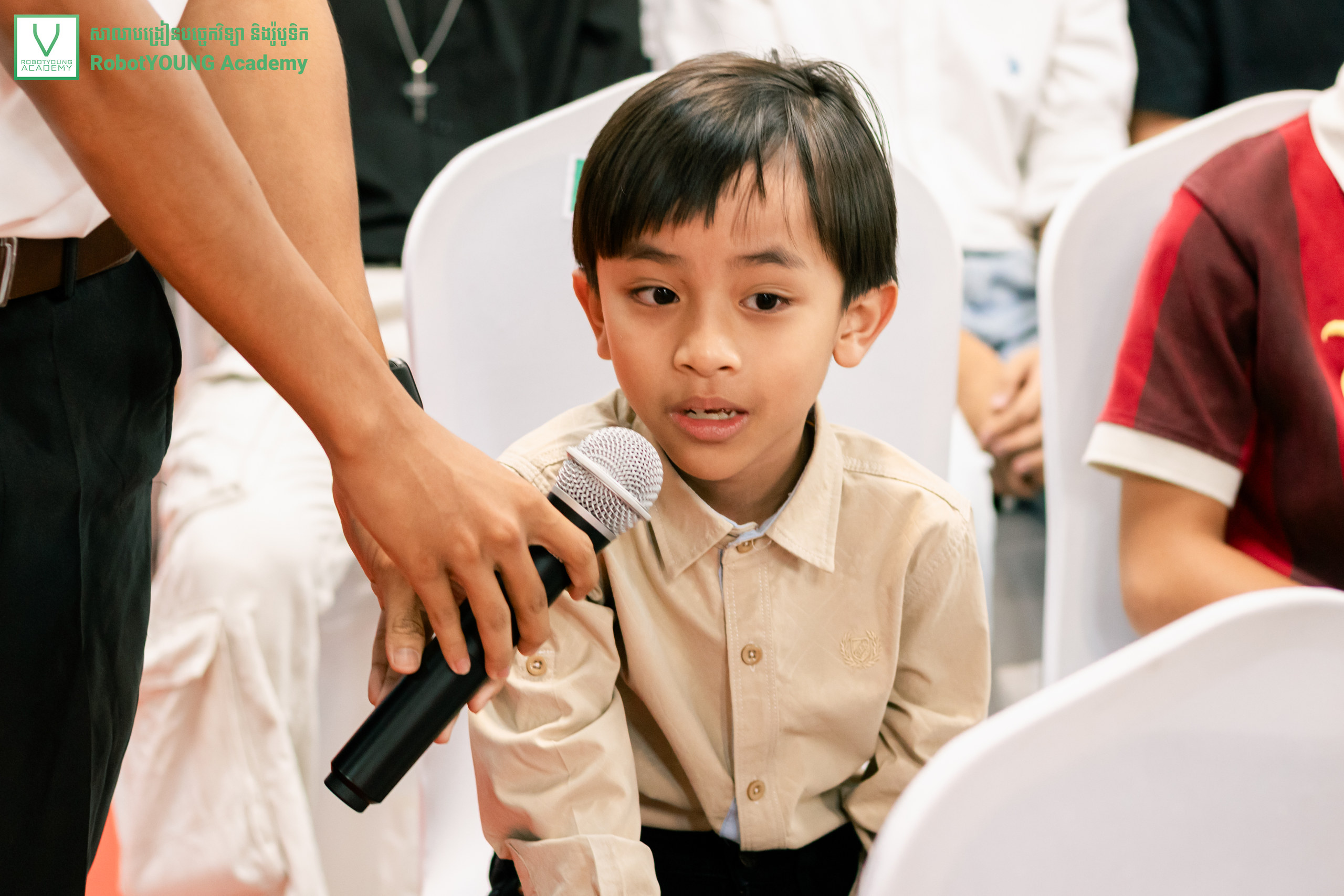 A child speaking into a microphone held by an adult among seated attendees