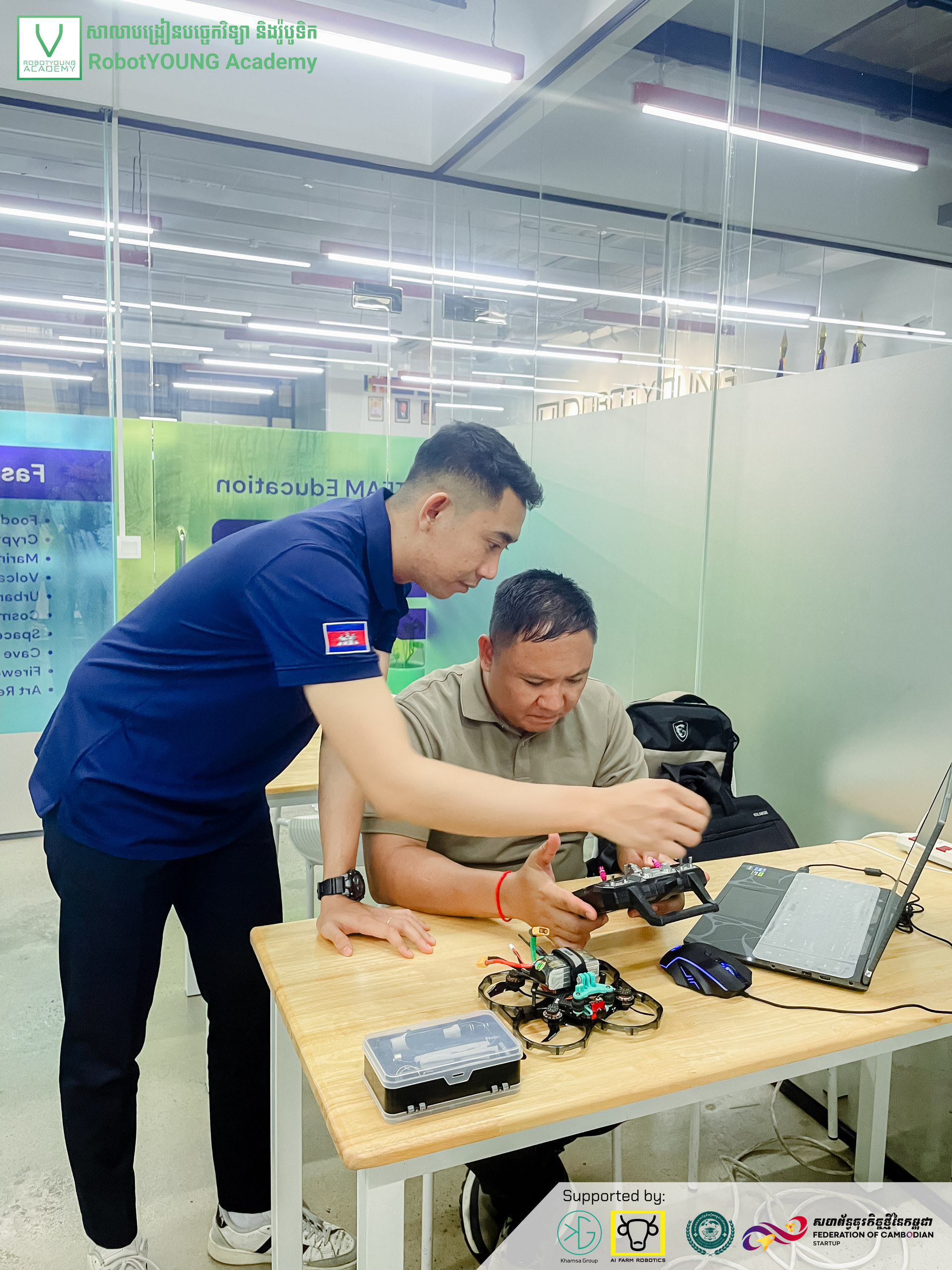 An instructor pointing out a robot component to a learner seated beside a laptop