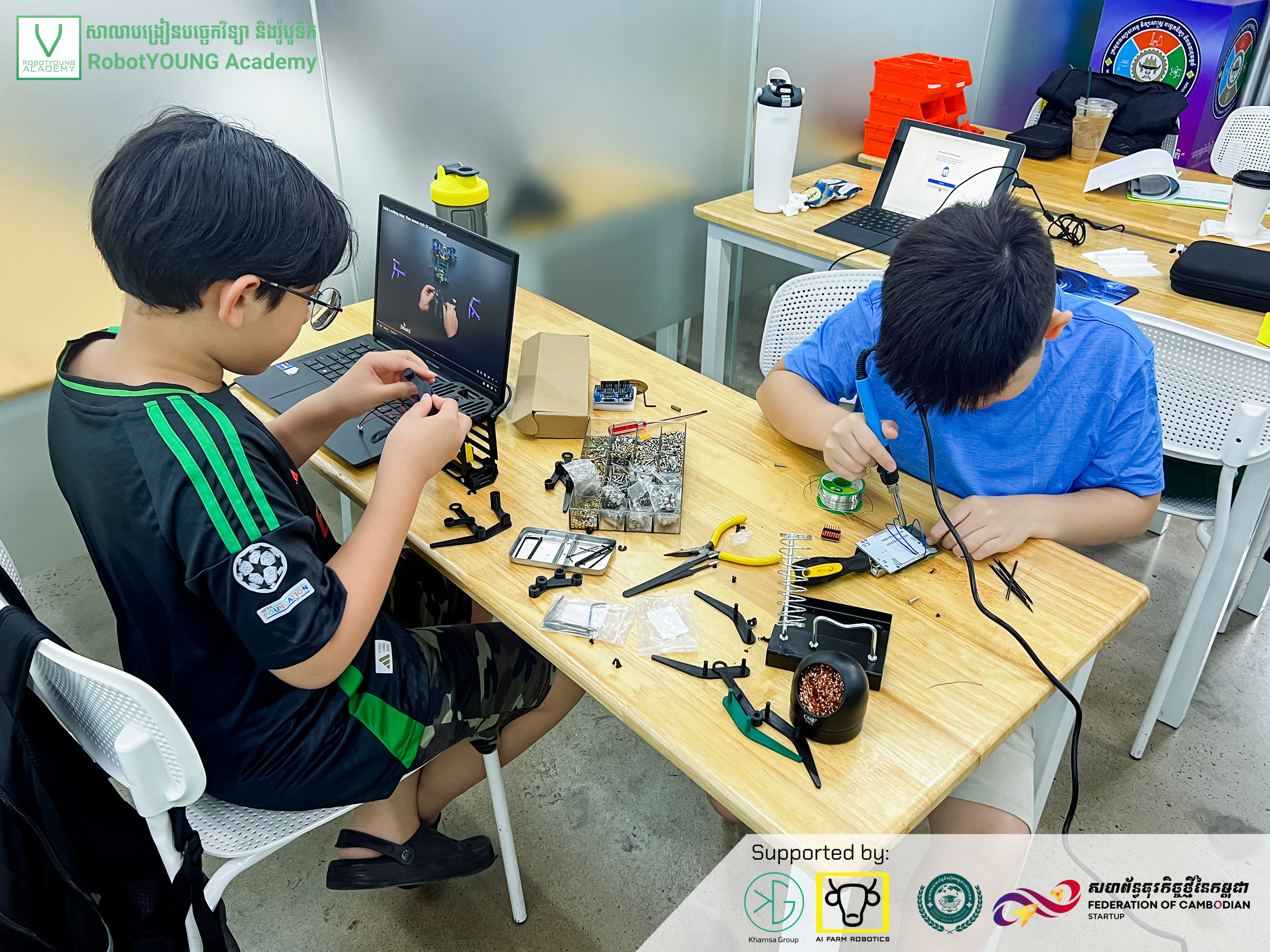 Illustrative academy photo of two students assembling electronics at a table with tools and a laptop