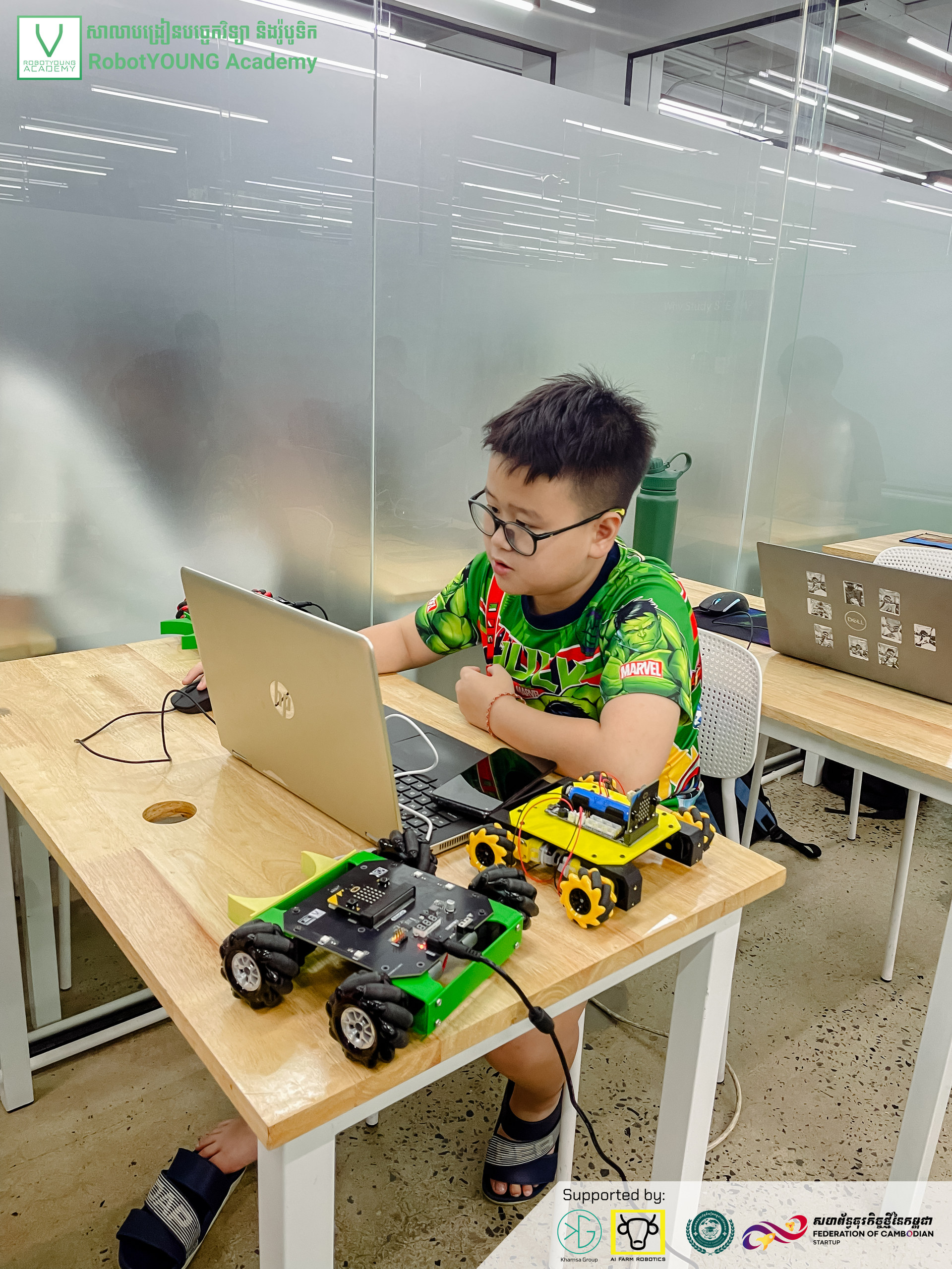 A student wearing glasses using a laptop beside green and yellow wheeled robots