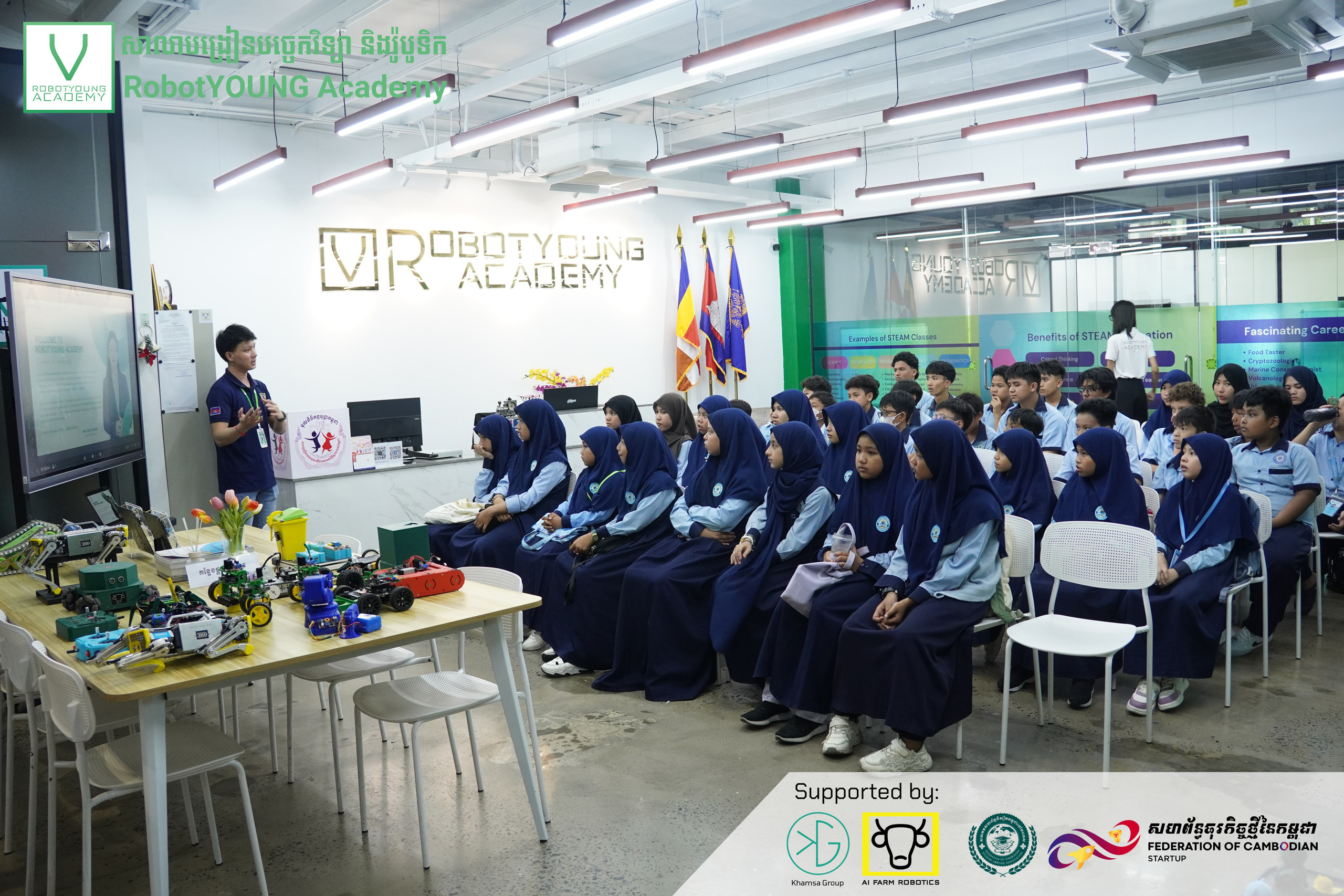 Students seated for an academy presentation beside a table of robot models
