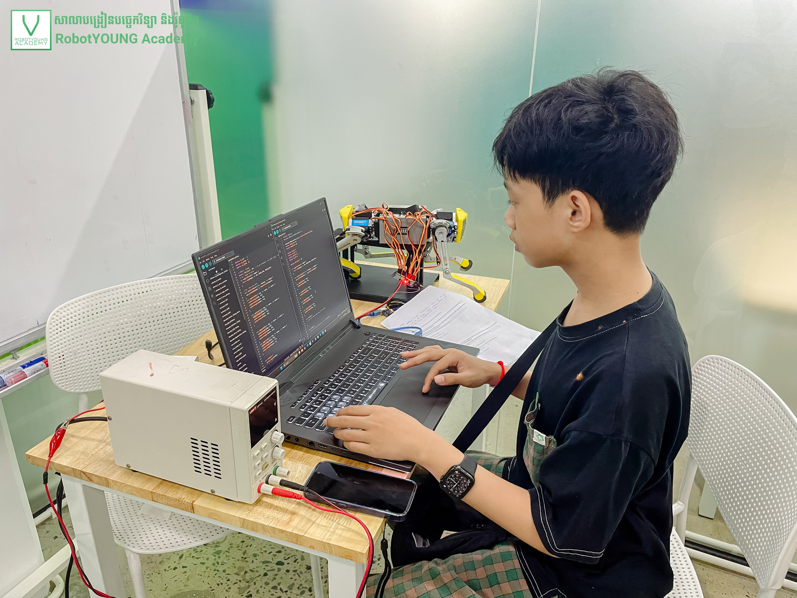 A student writing code on a laptop beside a robot