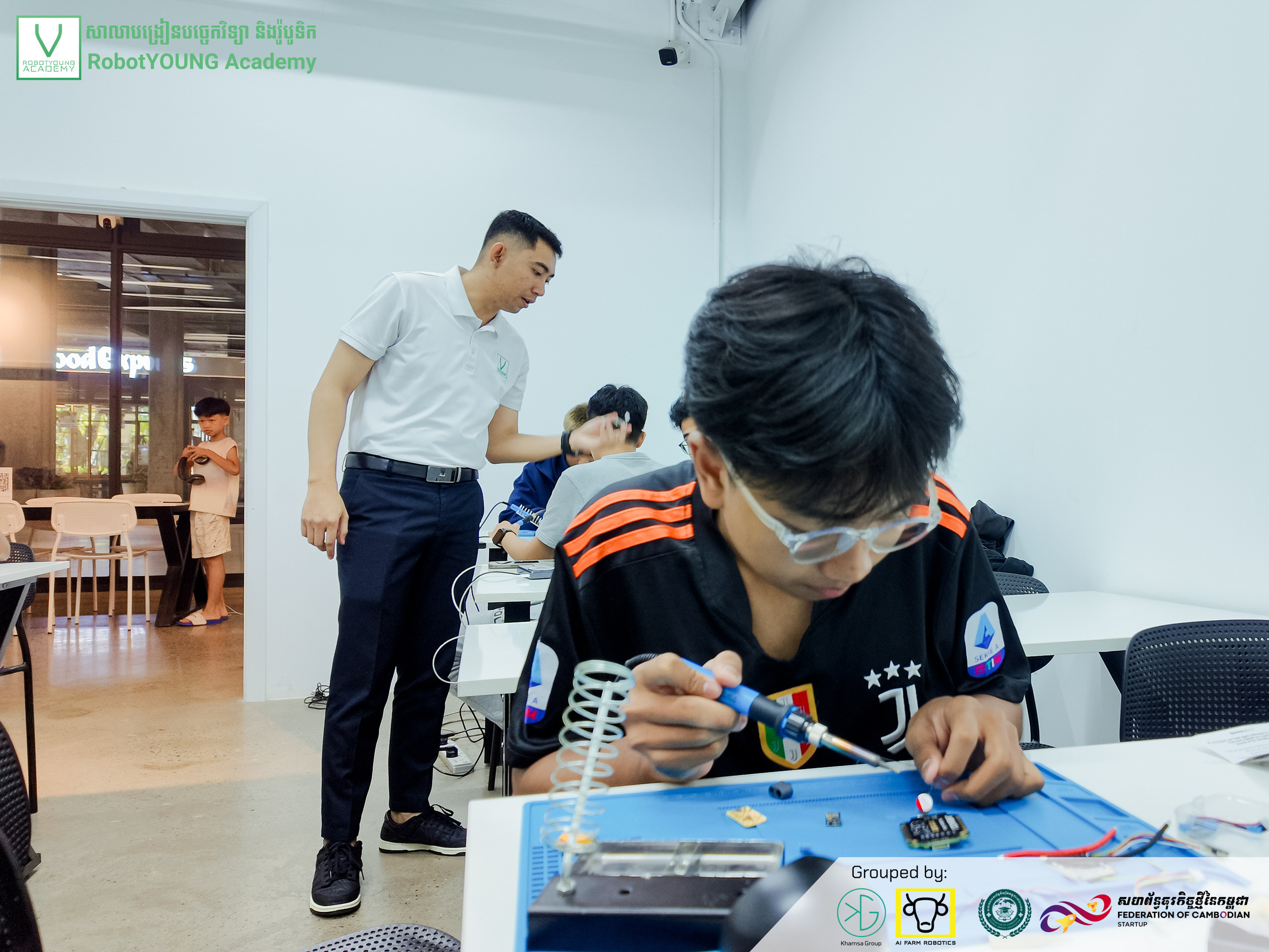 A learner concentrating on a circuit board at a blue work mat while an instructor stands nearby